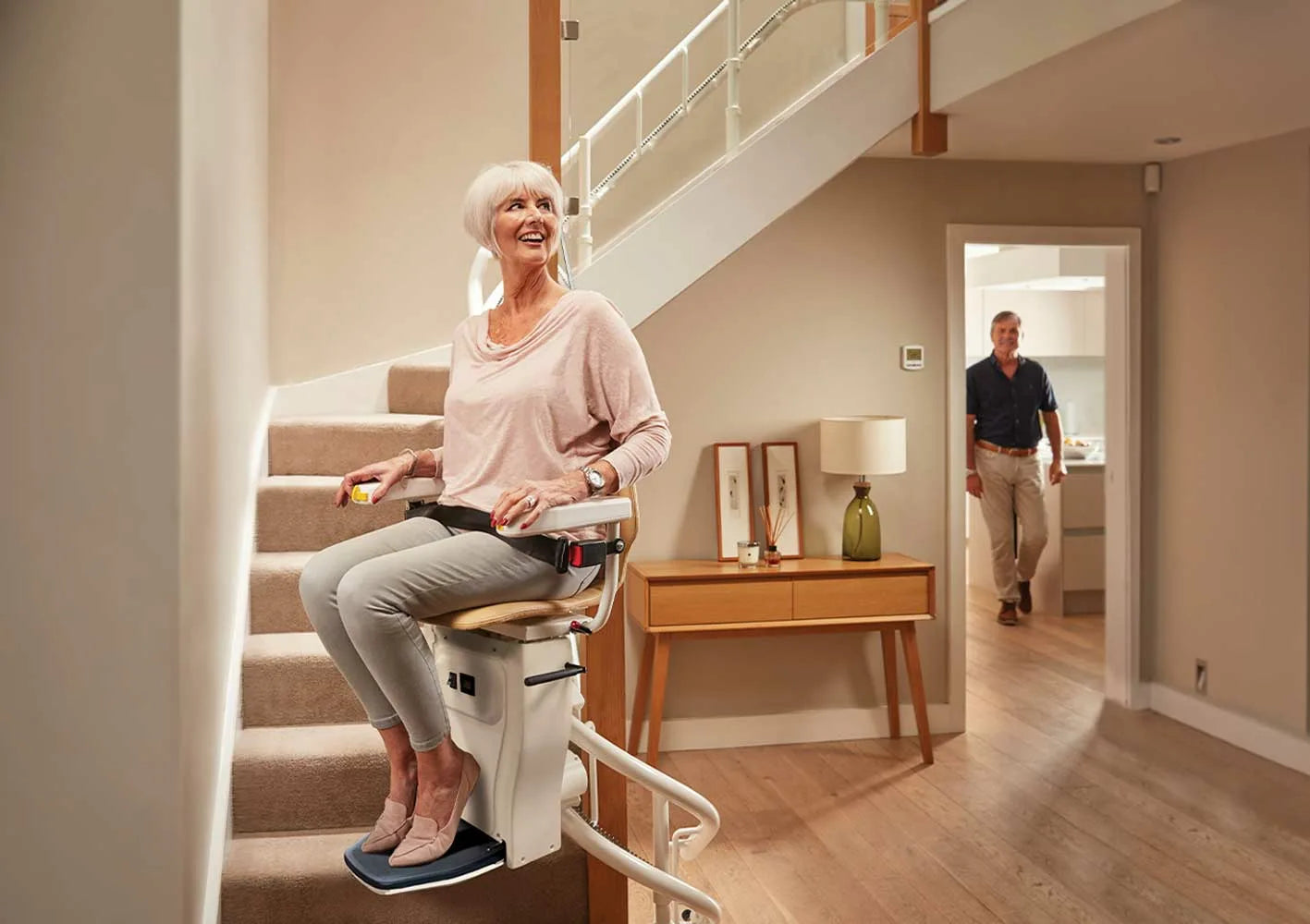 Woman using a stairlift in a home setting with a man in the background.